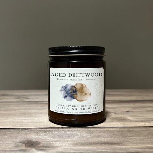 A jar candle with a label that reads 'AGED DRIFTWOOD', with a background featuring a small illustration of a wave and some driftwood on a beach.