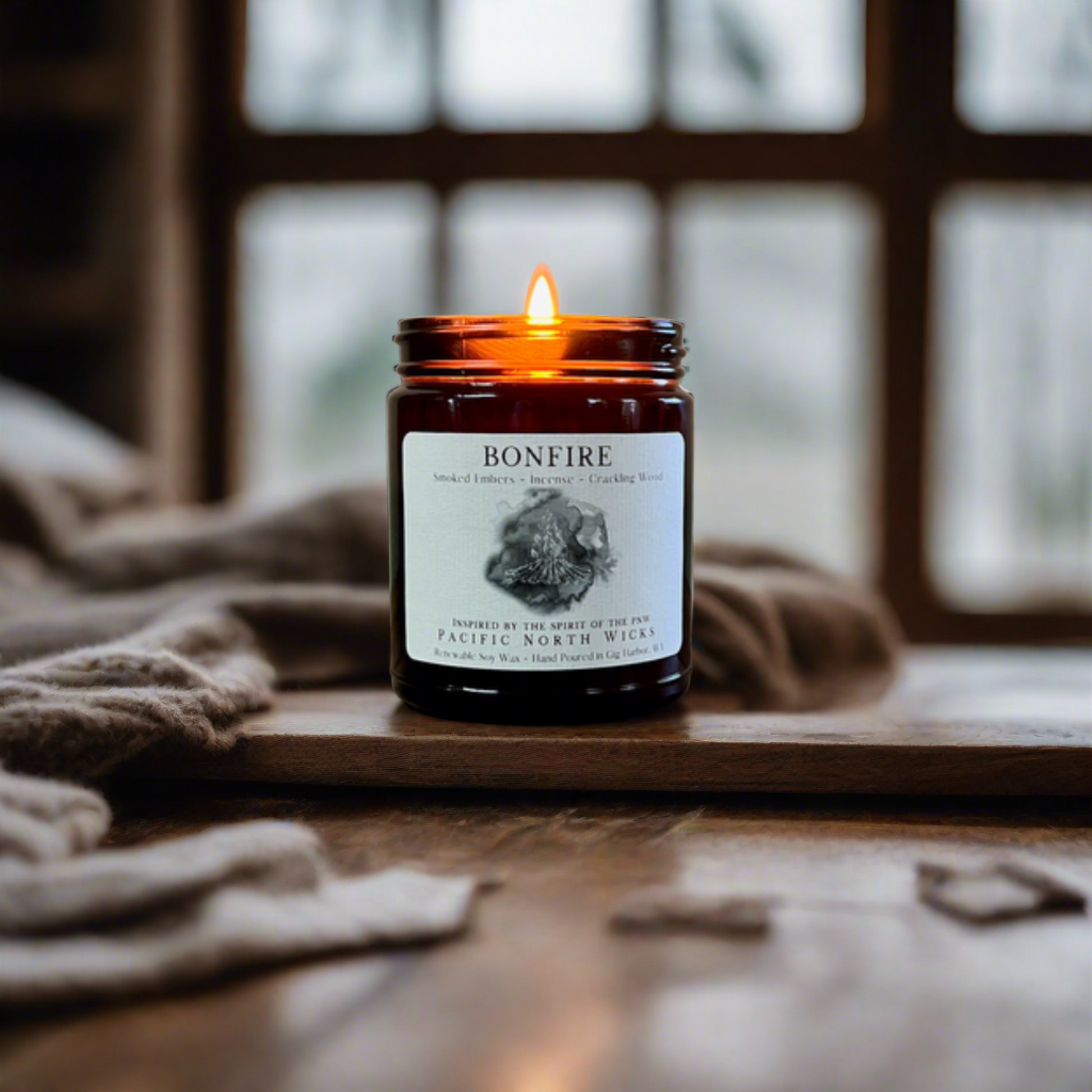 Firewood Scented Candle with notes of smoked embers, incense, and crackling wood.  Candle lit in Cozy Mountain Cabin with snow covered trees outside the window.