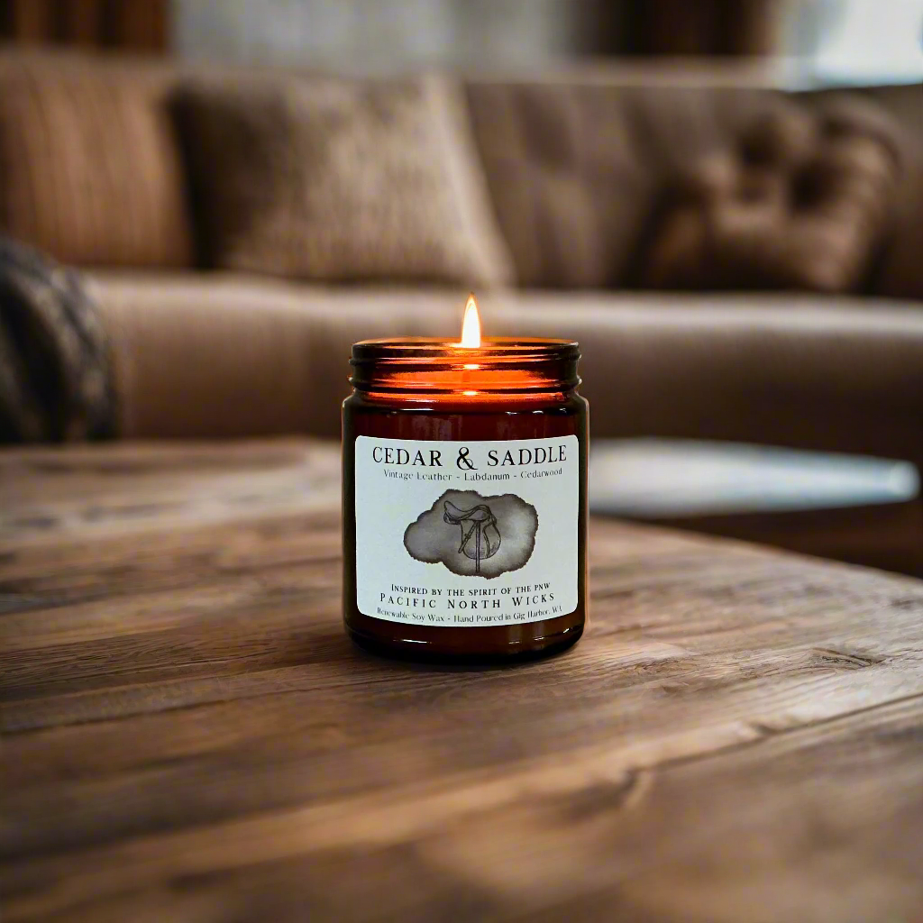 Candle in an amber jar labeled 'Cedar & Saddle' on a wooden coffee table with a blurred rustic living room background.
