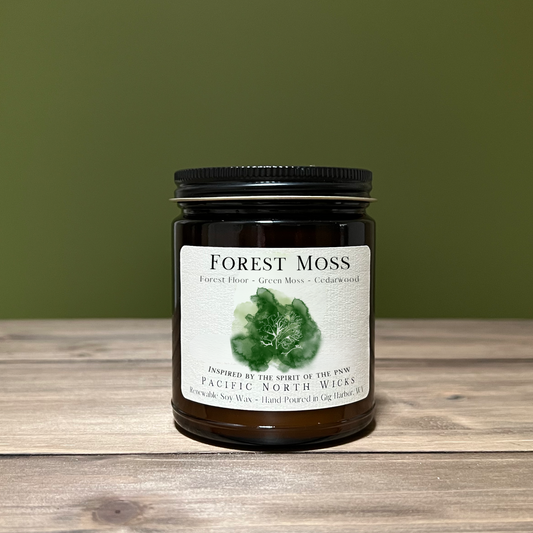 An amber jar soy candle with a label reading 'FOREST MOSS', placed on a wooden surface with a green background.