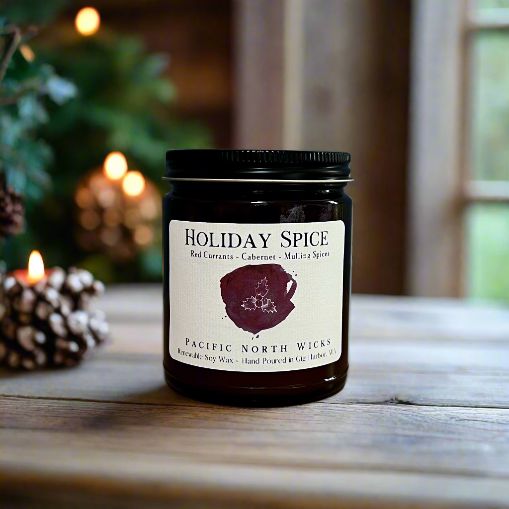 Holiday candle with Holiday Spice label with scent notes of red currants, cabernet, and mulling spices. Christmas tree and pine cones in blurred background.
