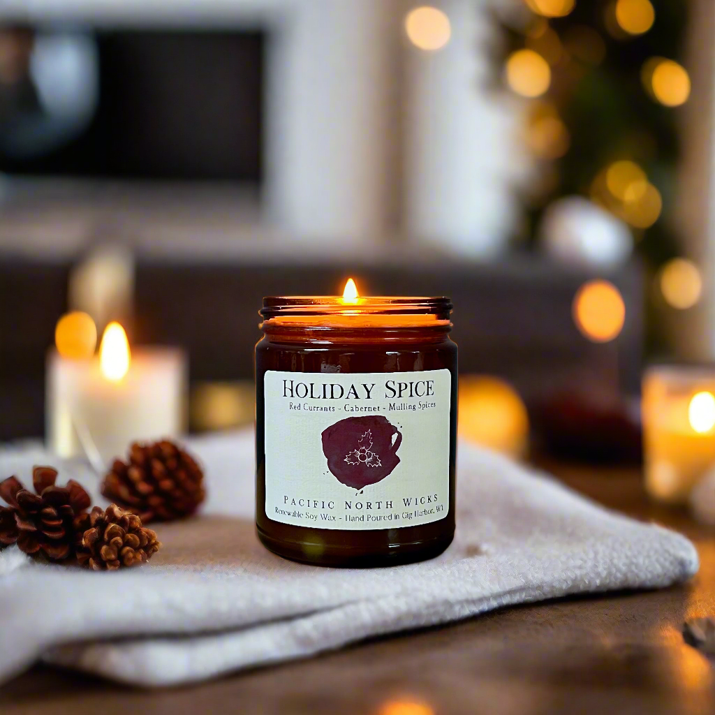 Christmas candle called Holiday Spice in amber jar burning in a cozy holiday setting with blurred lights in background.