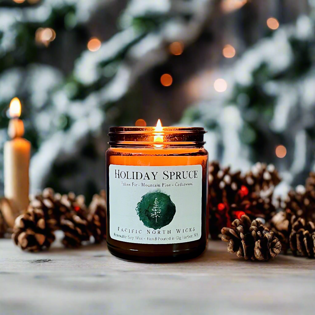 Holiday Spruce soy wax candle with flocked Christmas tree in background surrounded by pine cones