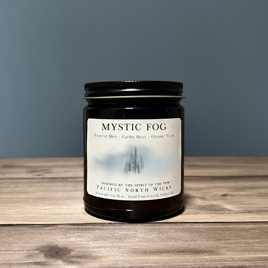 A black jar candle with a label that reads 'Mystic Fog', describing the fragrance as 'Forest Mist - Earthy Moss - Ozonic Veil'. The candle is placed on a wooden surface with a grey background.