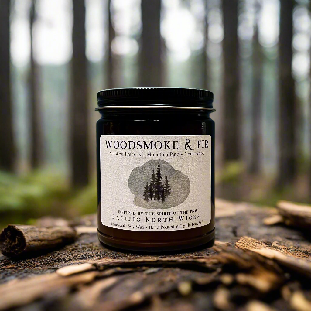 Woodsmoke & Fir Candle sitting on rustic bark in PNW woodsy setting with forest trees in background.