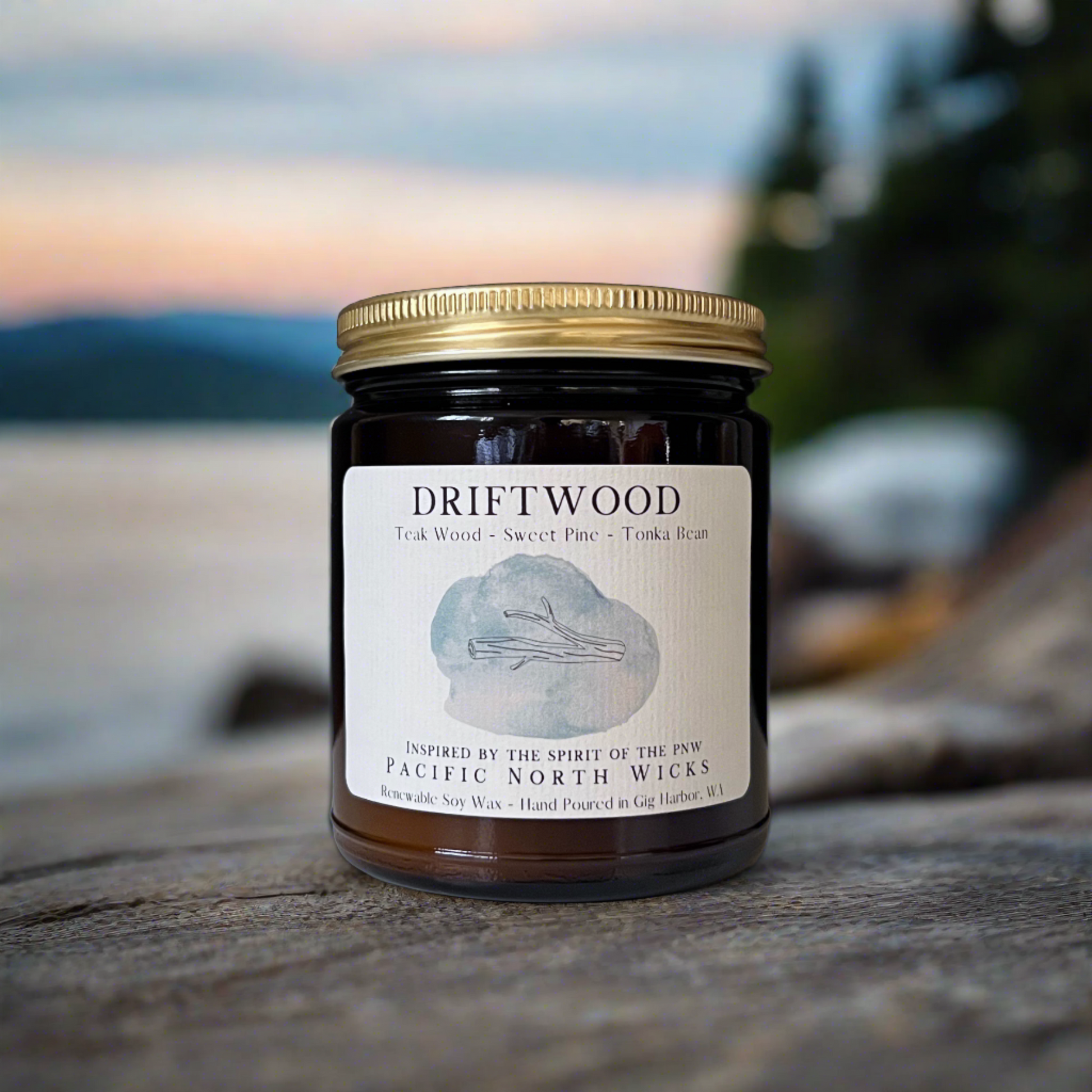 Driftwood Candle with gold lid on shores of Puget Sound with Salish Sea and forest trees in background.  Set on rustic driftwood at sunset.