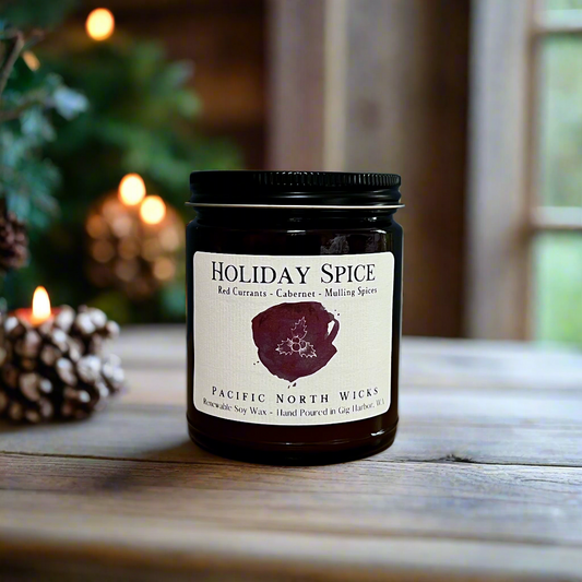 Holiday candle with Holiday Spice label with scent notes of red currants, cabernet, and mulling spices.  Christmas tree and pine cones in blurred background.